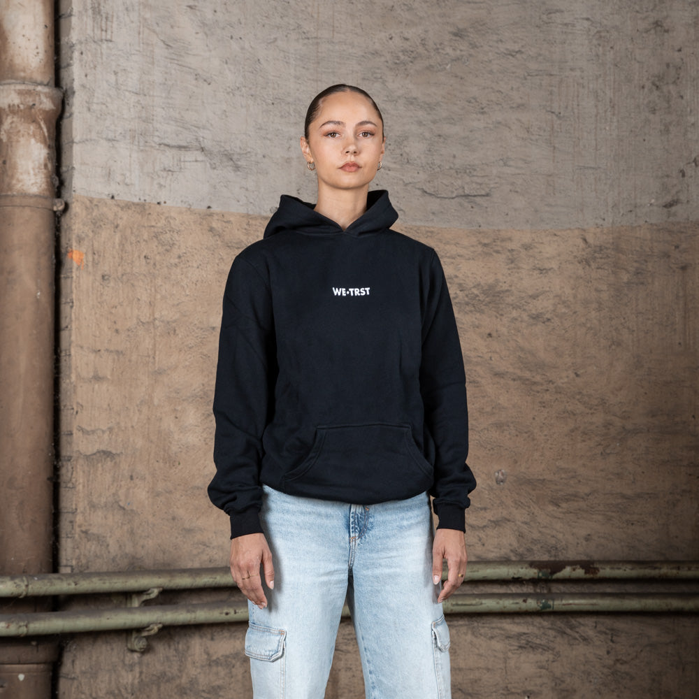 A female model in a black WE•TRST hoodie with white branding on the chest, styled with light-wash cargo jeans, standing against a raw concrete background.