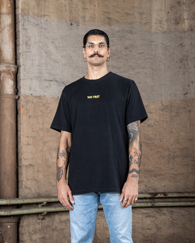 Ein männliches Modell, das ein schwarzes WE•TRST-T-Shirt mit einem gelben Logo auf der Brust trägt und dazu eine hellblaue Jeans, posiert vor einer Wand im Industriestil.