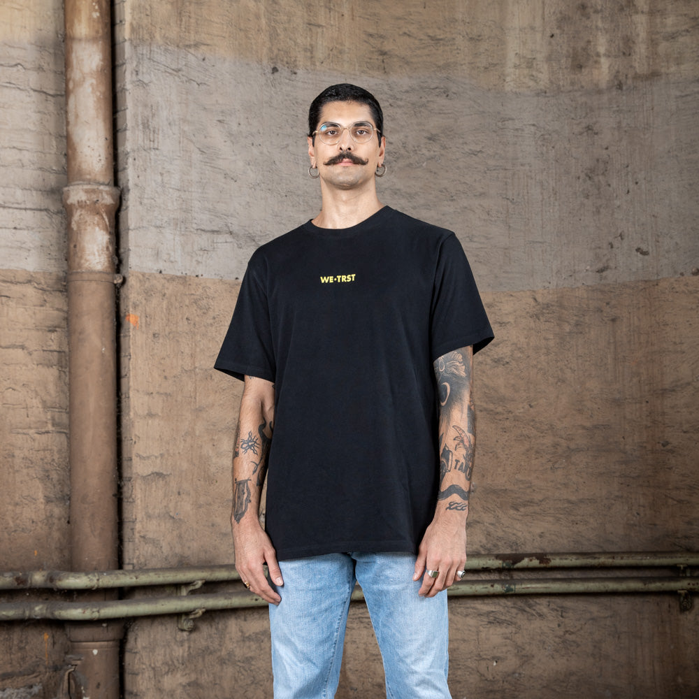 Ein männliches Modell, das ein schwarzes WE•TRST-T-Shirt mit einem gelben Logo auf der Brust trägt und dazu eine hellblaue Jeans, posiert vor einer Wand im Industriestil.