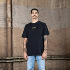 Ein männliches Modell, das ein schwarzes WE•TRST-T-Shirt mit einem gelben Logo auf der Brust trägt und dazu eine hellblaue Jeans, posiert vor einer Wand im Industriestil.