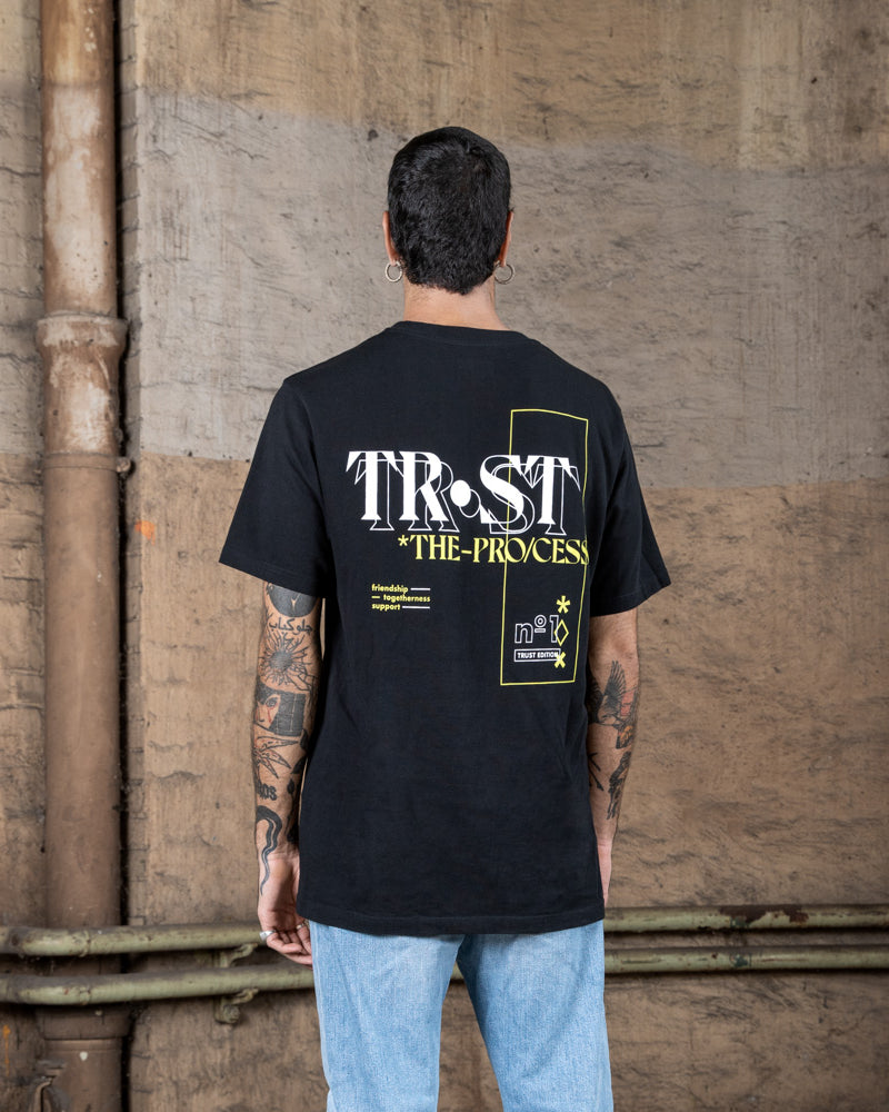 Rückenansicht eines männlichen Modells, das ein schwarzes WE•TRST-T-Shirt mit dem Schriftzug „TRST THE PROCESS“ und grafischen Details in Weiß und Gelb trägt.