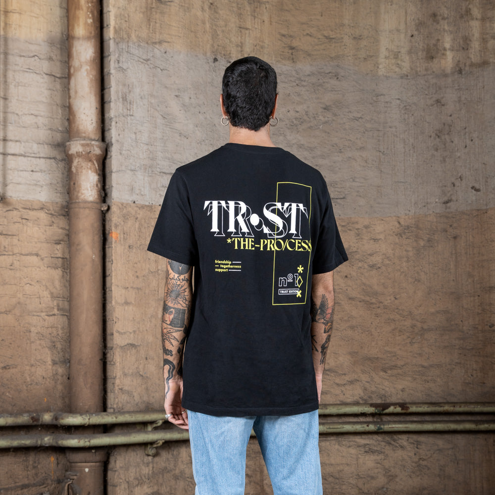 Back view of a male model wearing a black WE•TRST t-shirt with bold "TRST THE PROCESS" typography and graphic details in white and yellow.