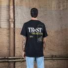 Rückenansicht eines männlichen Modells, das ein schwarzes WE•TRST-T-Shirt mit dem Schriftzug „TRST THE PROCESS“ und grafischen Details in Weiß und Gelb trägt.