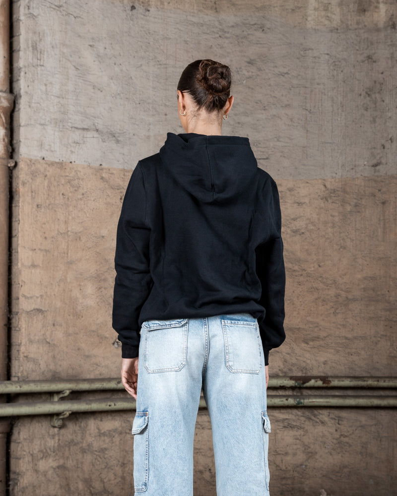 Rückenansicht eines weiblichen Models in einem schwarzen WE•TRST-Kapuzenpullover, der ein minimalistisches Design mit Kapuze und ohne zusätzliche Aufdrucke zeigt.