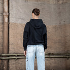 Rückenansicht eines weiblichen Models in einem schwarzen WE•TRST-Kapuzenpullover, der ein minimalistisches Design mit Kapuze und ohne zusätzliche Aufdrucke zeigt.