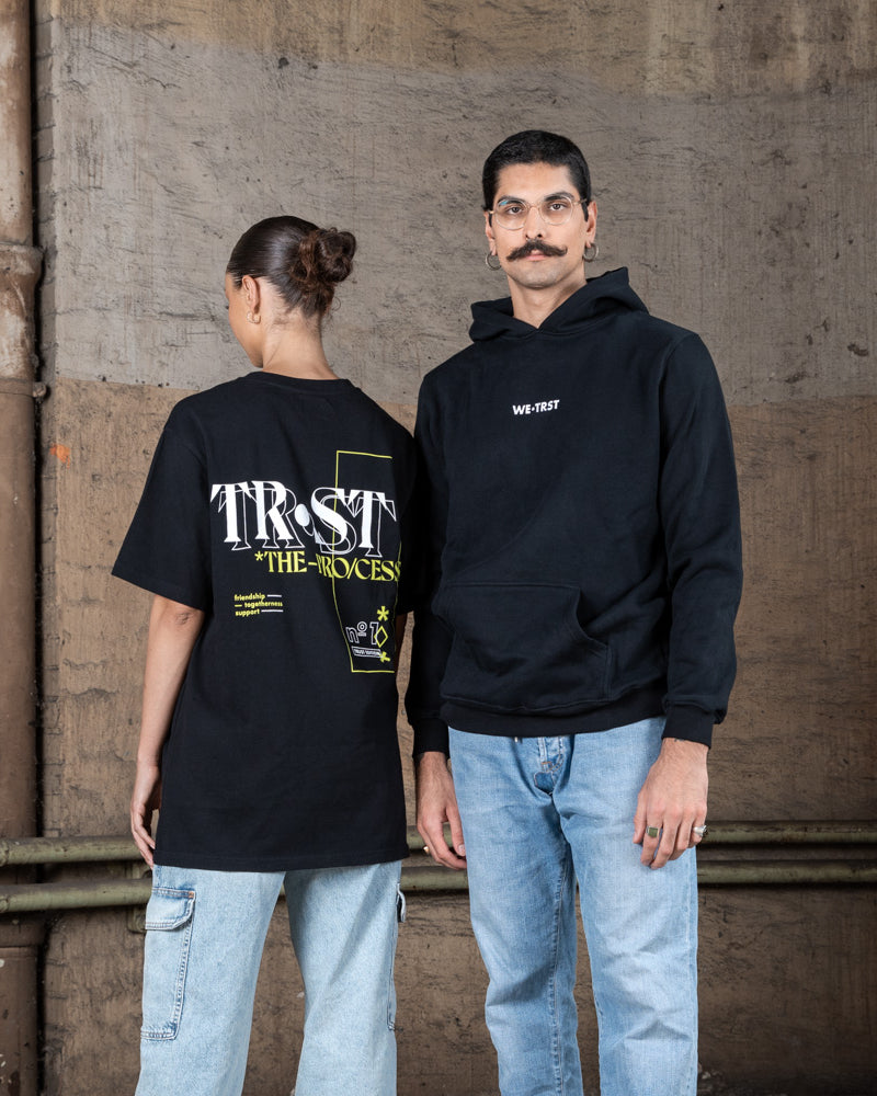 Zwei Models posieren zusammen in WE•TRST Streetwear. Die Frau trägt ein T-Shirt mit großem Rückenprint, der Mann einen schwarzen Hoodie mit dezentem Logo auf der Vorderseite.