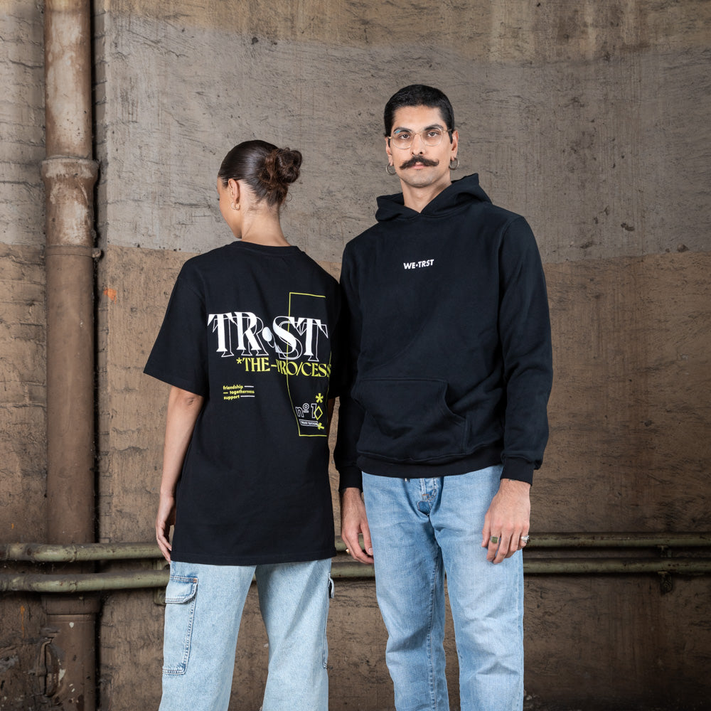 Zwei Models posieren zusammen in WE•TRST Streetwear. Die Frau trägt ein T-Shirt mit großem Rückenprint, der Mann einen schwarzen Hoodie mit dezentem Logo auf der Vorderseite.