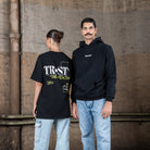 Zwei Models posieren zusammen in WE•TRST Streetwear. Die Frau trägt ein T-Shirt mit großem Rückenprint, der Mann einen schwarzen Hoodie mit dezentem Logo auf der Vorderseite.