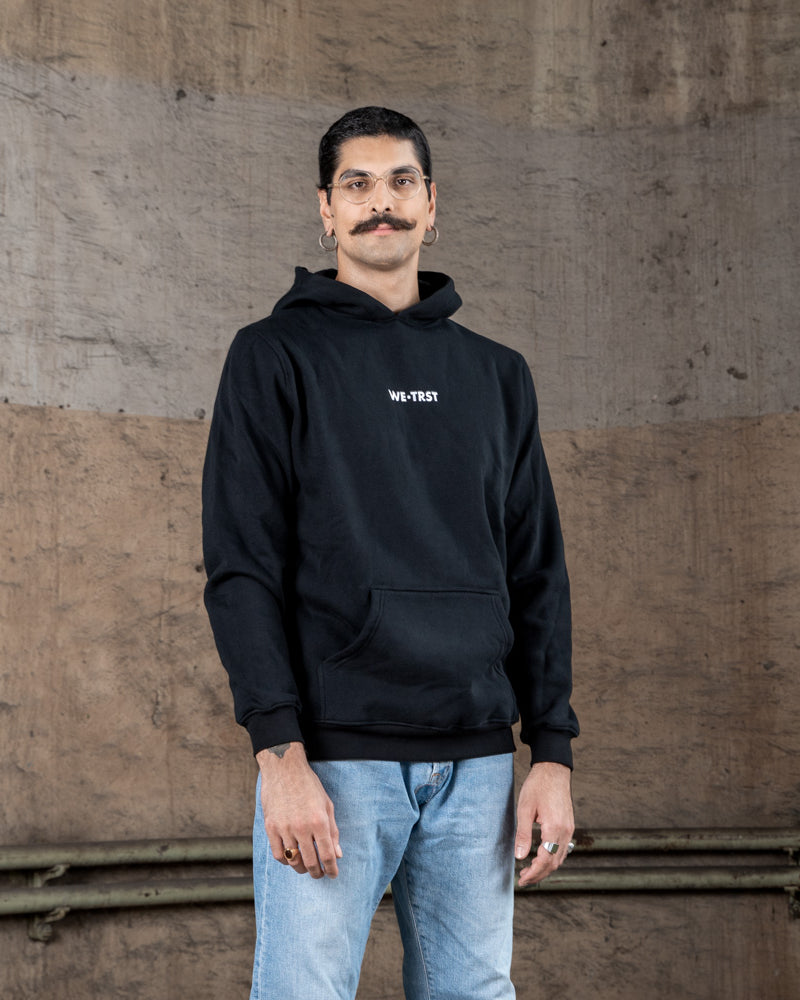 Ein männliches Model in einem schwarzen WE•TRST-Kapuzenpulli mit weißem Logo auf der Brust steht vor einem strukturierten Industriehintergrund.