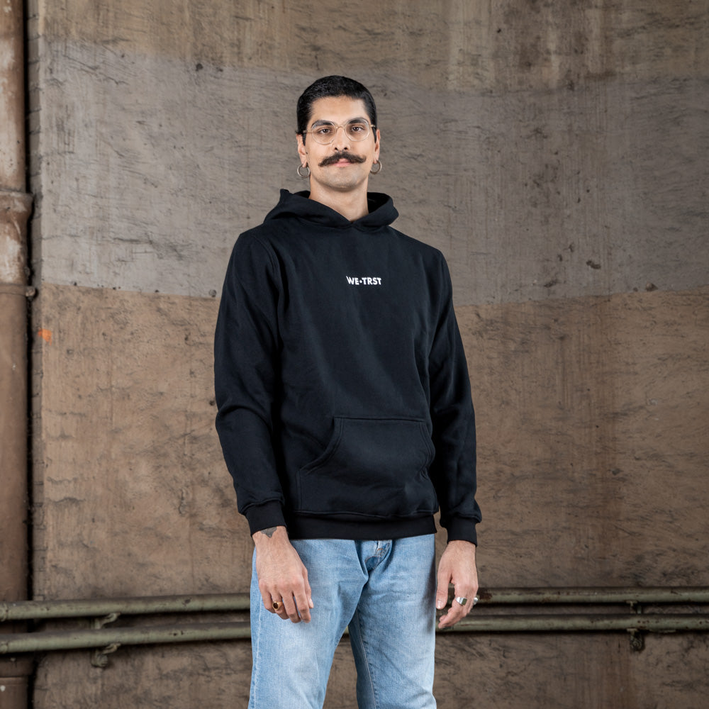 Ein männliches Model in einem schwarzen WE•TRST-Kapuzenpulli mit weißem Logo auf der Brust steht vor einem strukturierten Industriehintergrund.