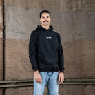 Ein männliches Model in einem schwarzen WE•TRST-Kapuzenpulli mit weißem Logo auf der Brust steht vor einem strukturierten Industriehintergrund.