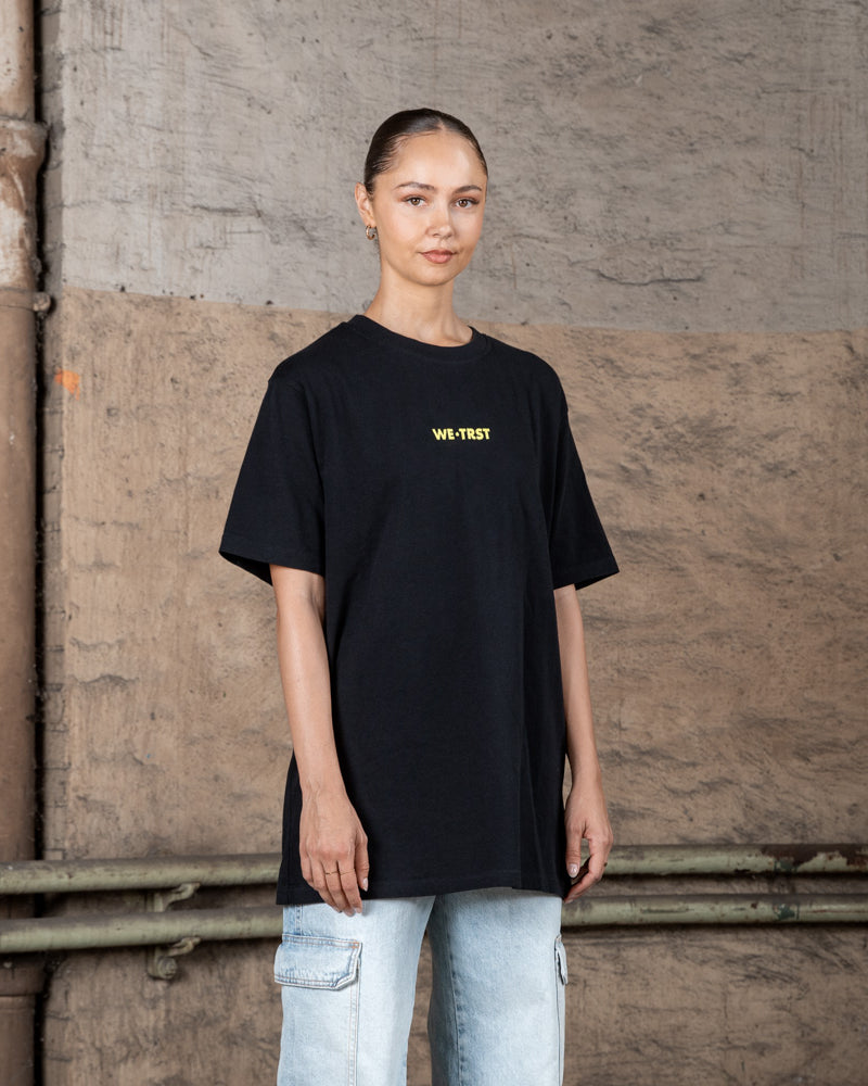 Ein weibliches Modell in einem schwarzen WE•TRST T-Shirt in Übergröße mit gelbem Logo, gepaart mit hellen Cargo-Jeans, steht in einer städtischen Industrieumgebung.
