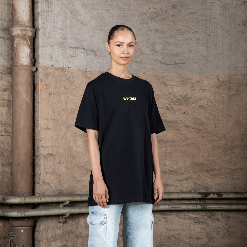 Ein weibliches Modell in einem schwarzen WE•TRST T-Shirt in Übergröße mit gelbem Logo, gepaart mit hellen Cargo-Jeans, steht in einer städtischen Industrieumgebung.