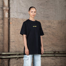 Ein weibliches Modell in einem schwarzen WE•TRST T-Shirt in Übergröße mit gelbem Logo, gepaart mit hellen Cargo-Jeans, steht in einer städtischen Industrieumgebung.