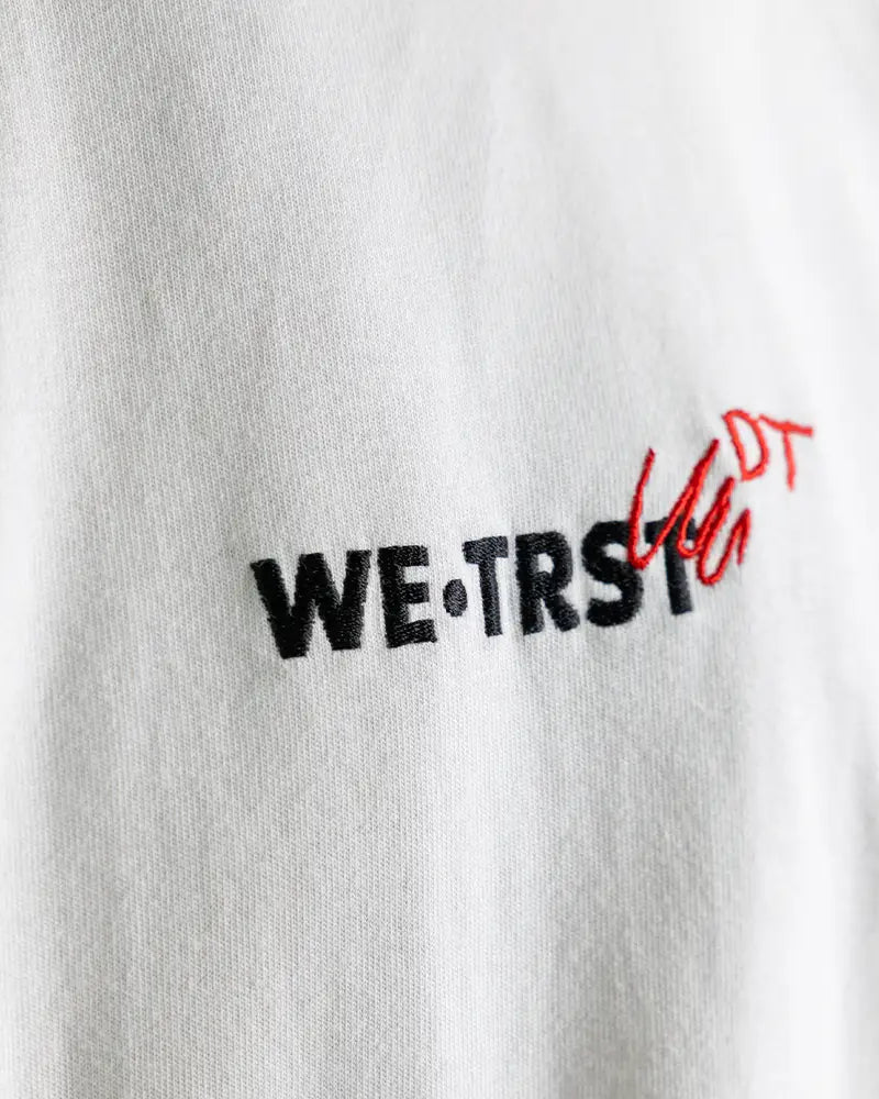 Nahaufnahme des WE•TRST Dangel Twins-Logos auf weissem Shirt – Stickerei mit rotem Doodle-Effekt.