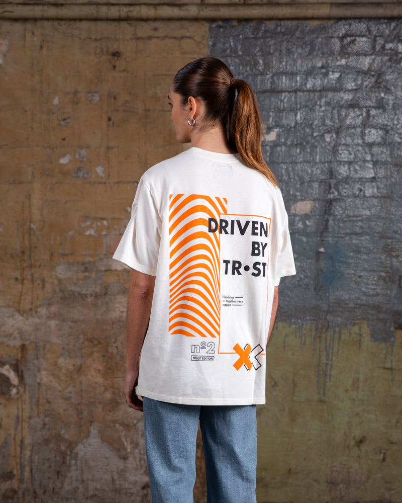 Rückenansicht eines weiblichen Models, das das WE•TRST „Driven by Trust“ T-Shirt in Übergröße trägt, wobei die auffällige orangefarbene Grafik und das typografische Design hervorgehoben werden.