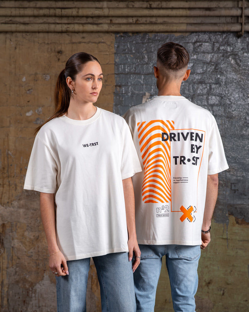Seite an Seite zeigen männliche und weibliche Models das Vorder- und Rückseitendesign des WE•TRST „Driven by Trust“ T-Shirts in Off-White.