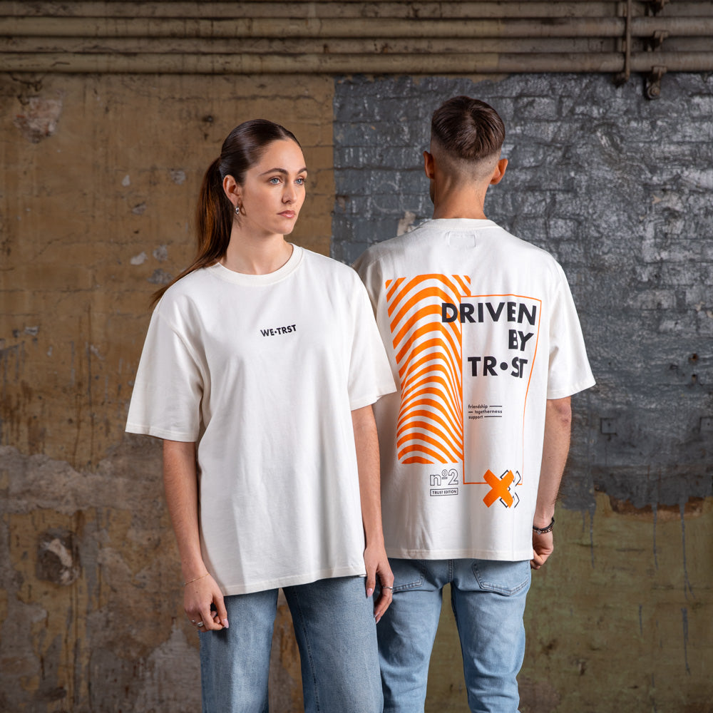 Seite an Seite zeigen männliche und weibliche Models das Vorder- und Rückseitendesign des WE•TRST „Driven by Trust“ T-Shirts in Off-White.