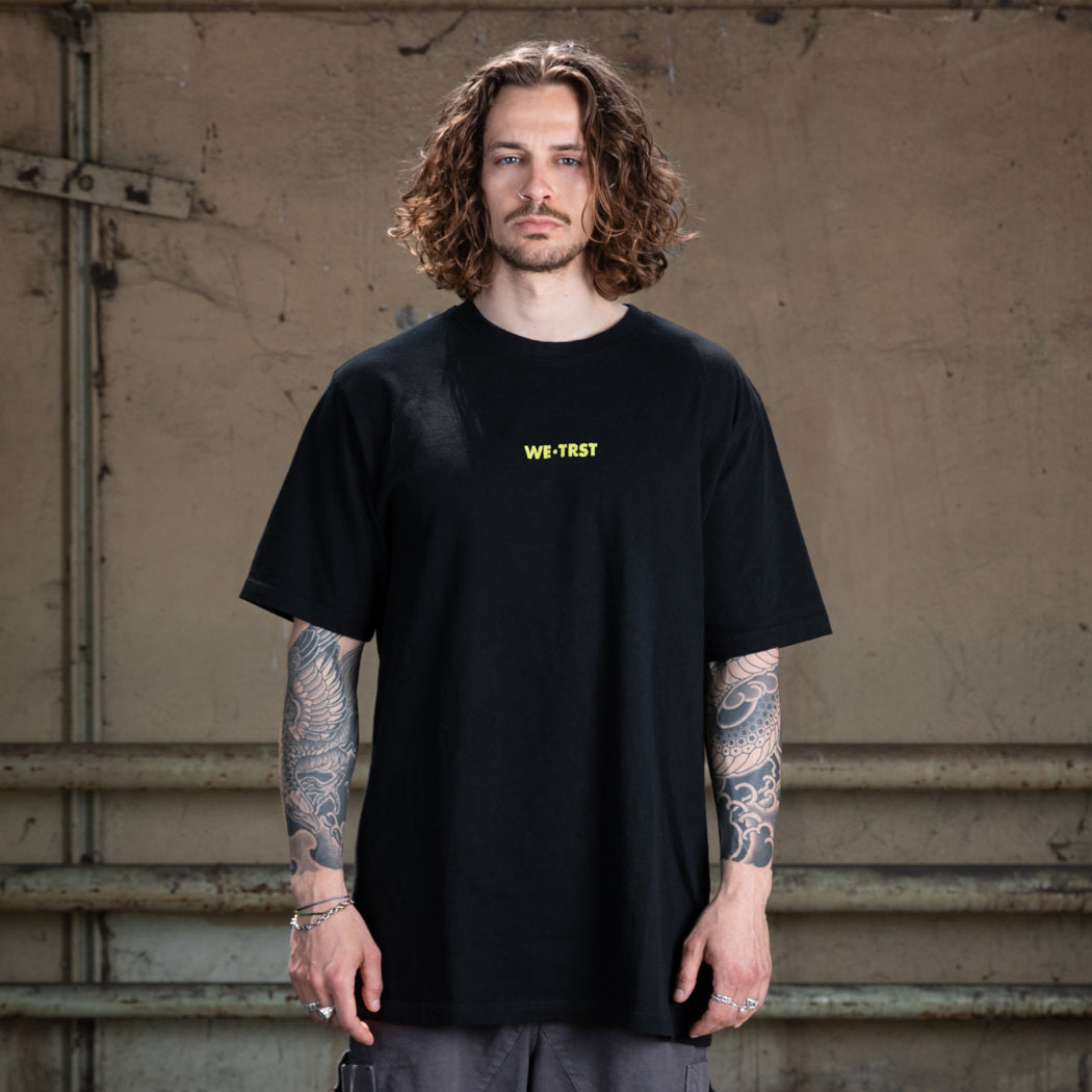 Ein männliches Modell mit gewelltem braunem Haar und Tätowierungen auf beiden Armen, das ein schwarzes WE•TRST-T-Shirt in Übergröße mit einem gelben Brustlogo trägt, steht vor einer Industriewand.