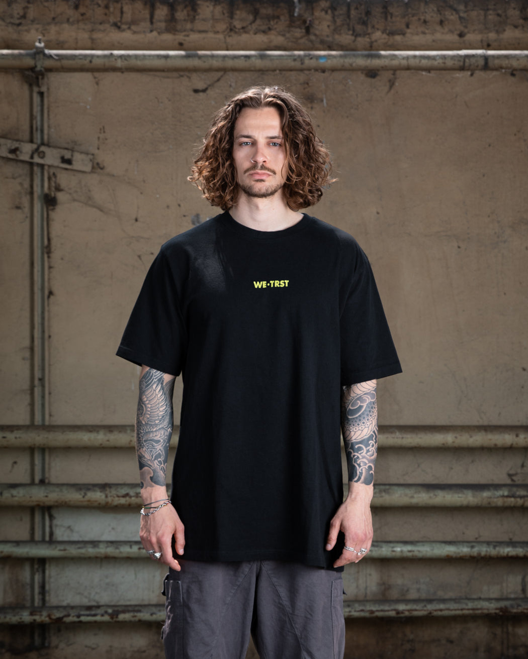Ein männliches Modell mit gewelltem braunem Haar und Tätowierungen auf beiden Armen, das ein schwarzes WE•TRST-T-Shirt in Übergröße mit einem gelben Brustlogo trägt, steht vor einer Industriewand.