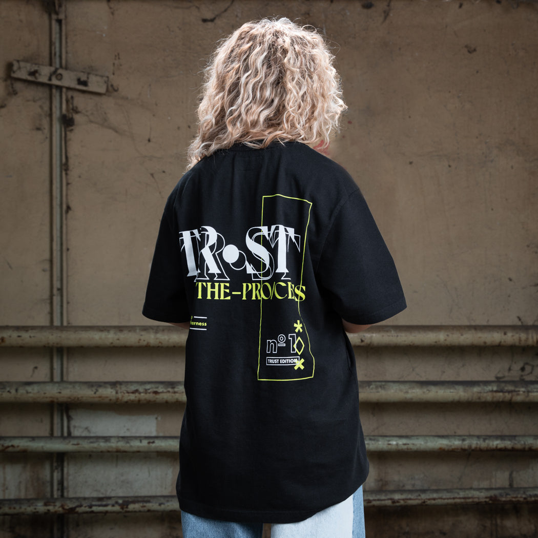 Rückenansicht eines weiblichen Modells mit blondem, lockigem Haar, das ein schwarzes WE•TRST-T-Shirt mit dem großen grafischen Aufdruck „TRST THE PROCESS“ in Weiß und Gelb trägt.