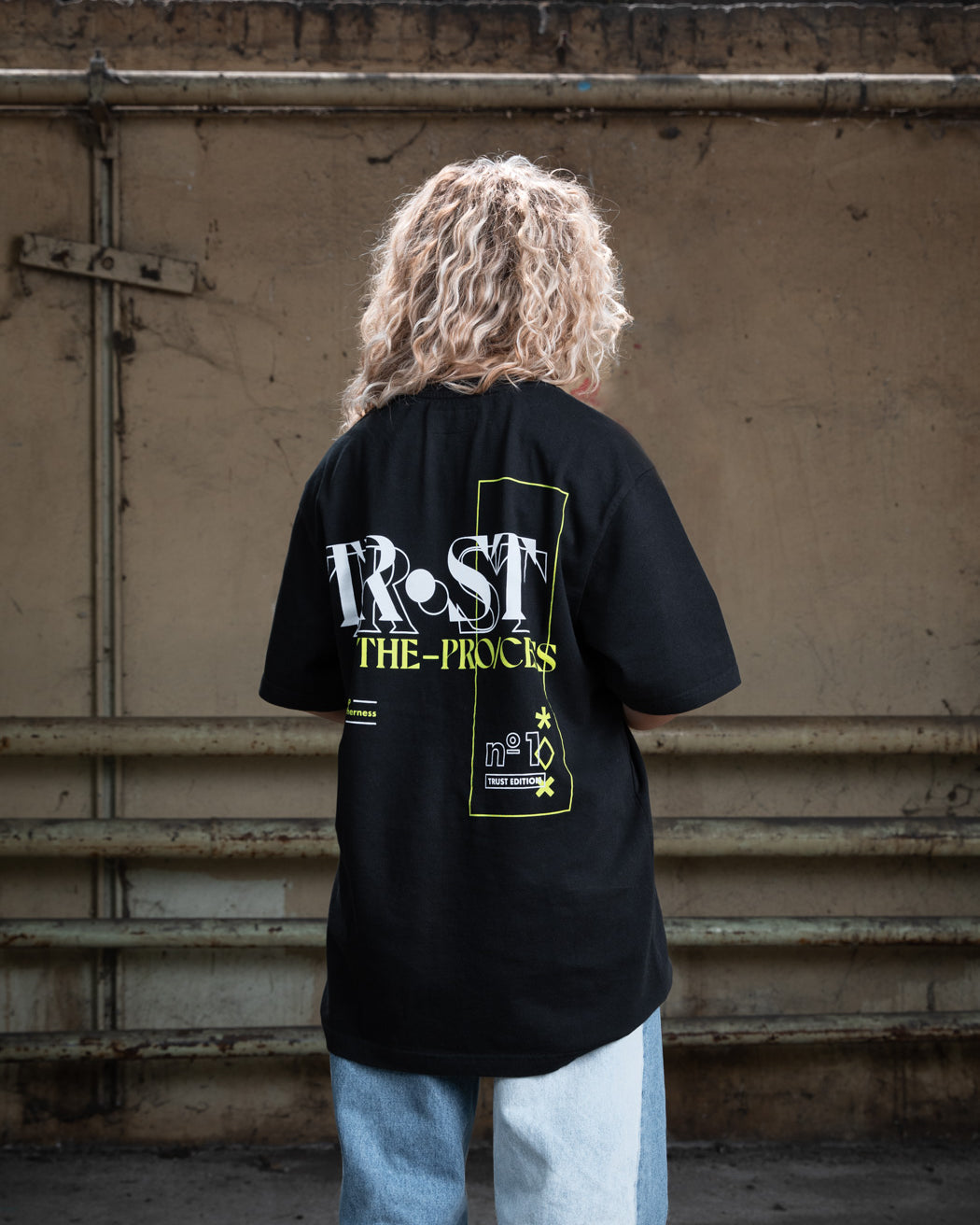 Rückenansicht eines weiblichen Modells mit blondem, lockigem Haar, das ein schwarzes WE•TRST-T-Shirt mit dem großen grafischen Aufdruck „TRST THE PROCESS“ in Weiß und Gelb trägt.