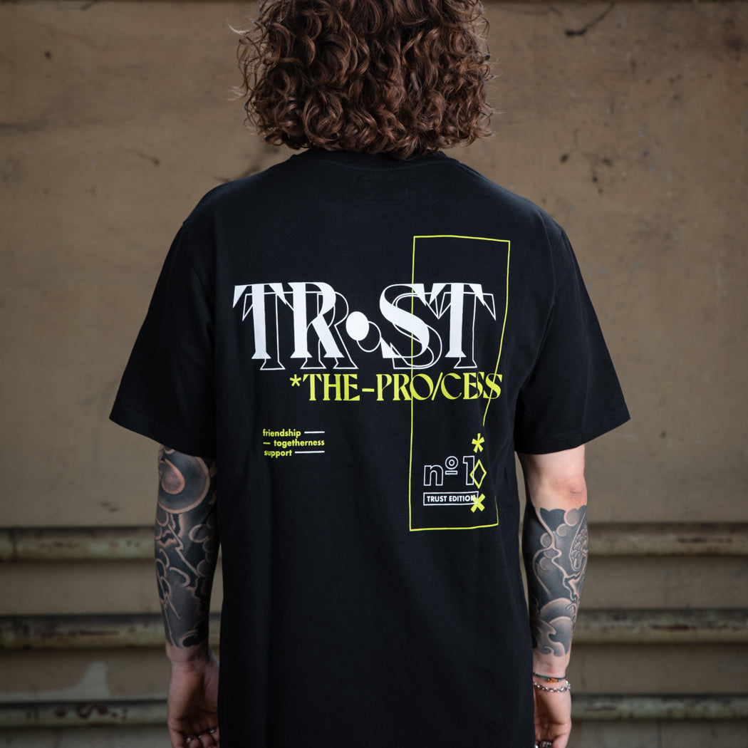 Nahaufnahme des Rückendesigns eines schwarzen WE•TRST-T-Shirts, das die Typografie „TRST THE PROCESS“ und grafische Elemente in Weiß und Neongelb zeigt.