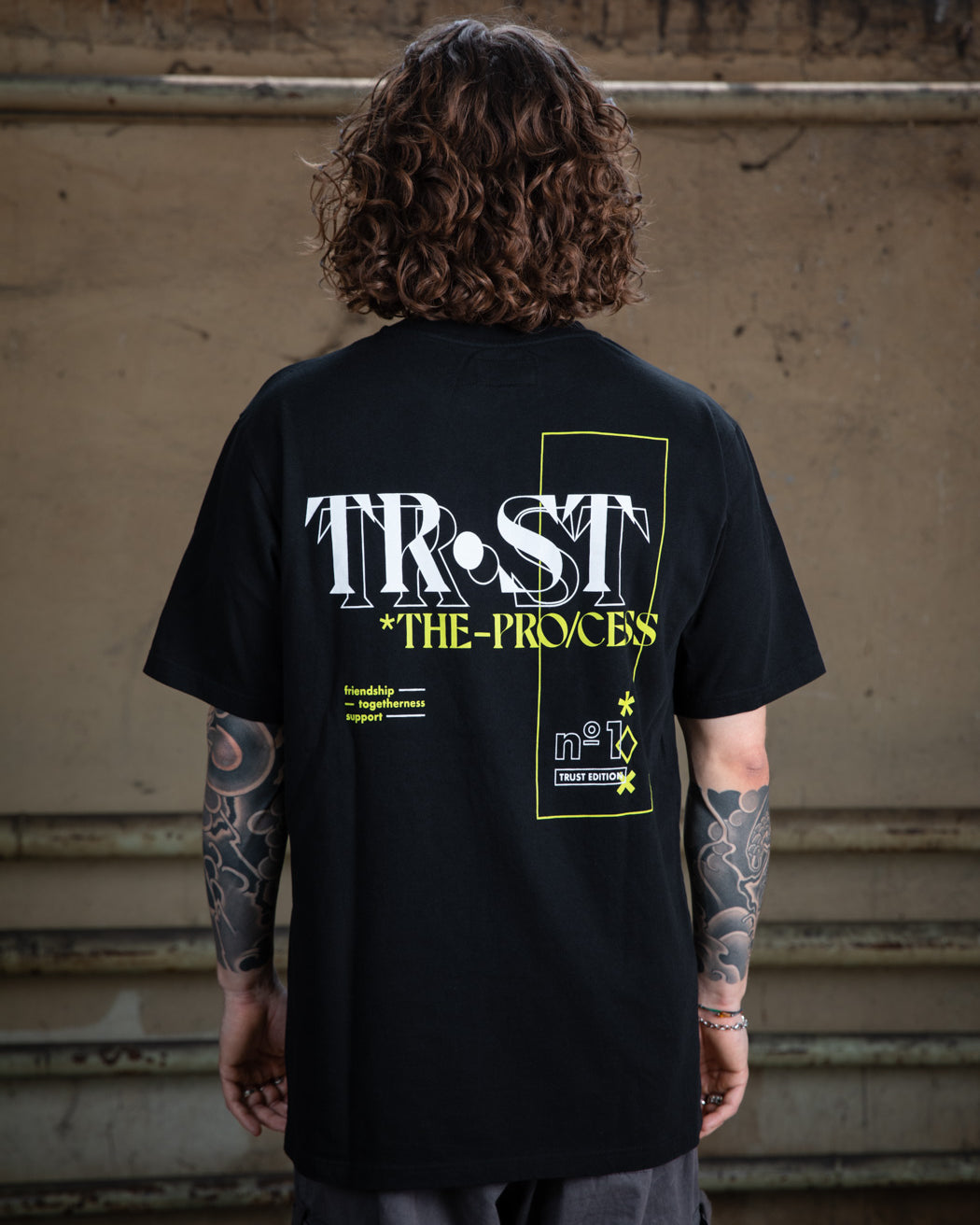 Nahaufnahme des Rückendesigns eines schwarzen WE•TRST-T-Shirts, das die Typografie „TRST THE PROCESS“ und grafische Elemente in Weiß und Neongelb zeigt.