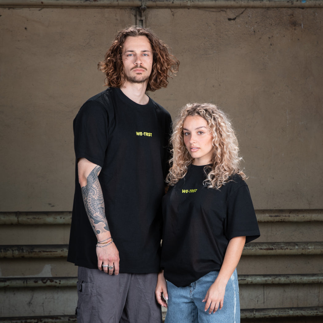 Ein männliches Model in einem schwarzen WE•TRST-T-Shirt mit Tätowierungen auf den Armen, gepaart mit einer Cargohose, steht vor einer rohen Betonwand.
