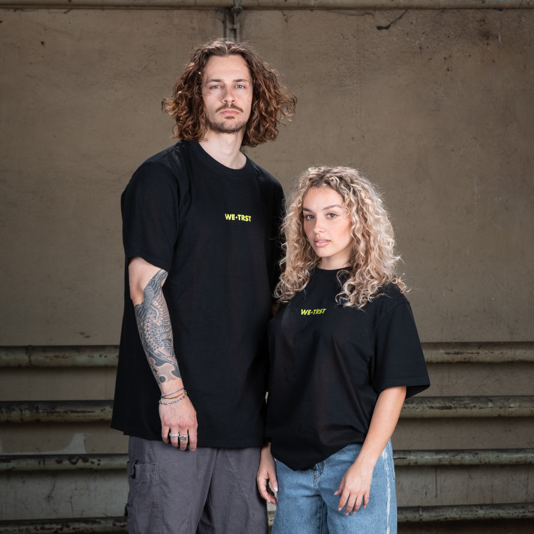 Ein männliches Model in einem schwarzen WE•TRST-T-Shirt mit Tätowierungen auf den Armen, gepaart mit einer Cargohose, steht vor einer rohen Betonwand.