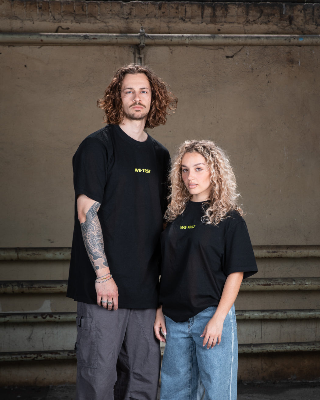 Ein männliches Model in einem schwarzen WE•TRST-T-Shirt mit Tätowierungen auf den Armen, gepaart mit einer Cargohose, steht vor einer rohen Betonwand.