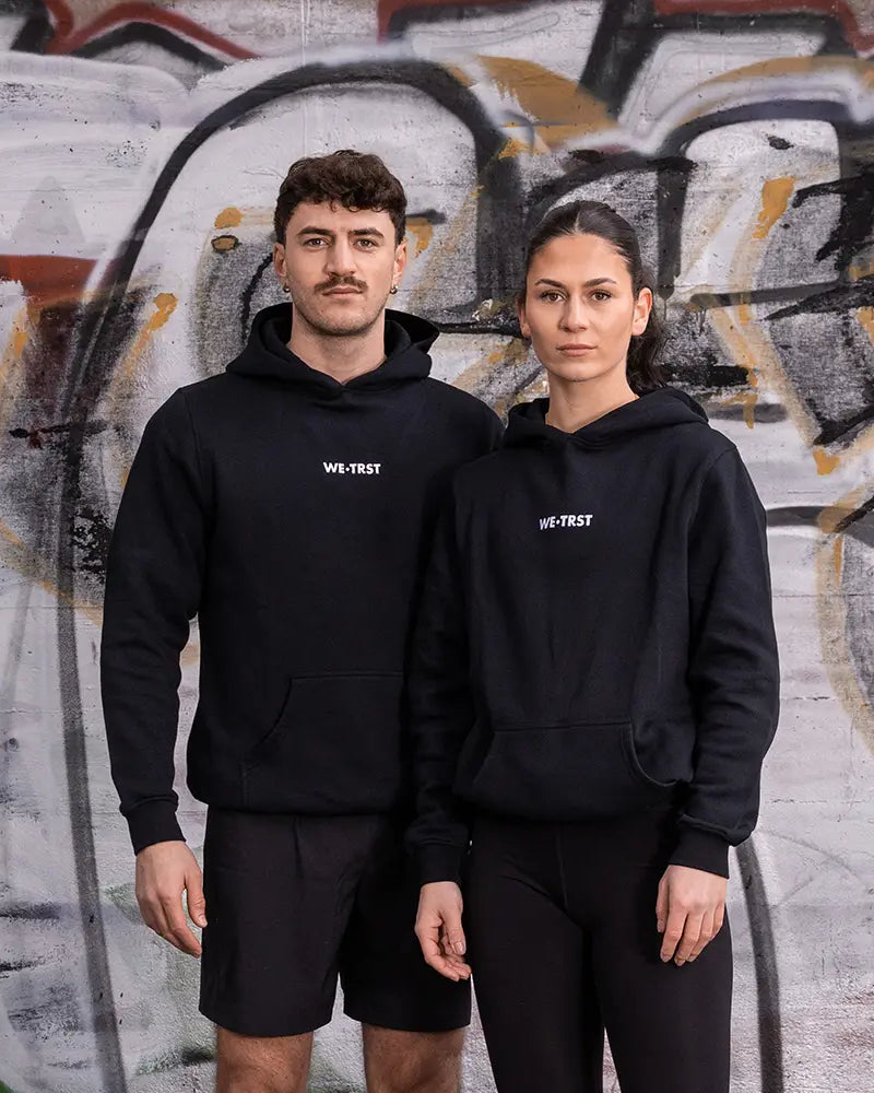 WE•TRST Running Department Hoodie – schwarze Unisex Streetwear für Läufer:innen vor urbaner Graffiti-Wand