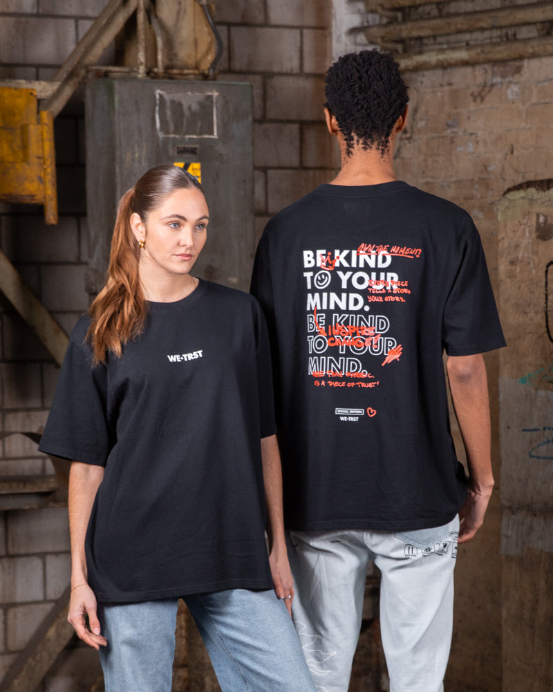 Zwei Models, die schwarze WE•TRST-T-Shirts in Übergröße mit einem Motiv für psychische Gesundheit tragen, posieren in einer städtischen Industriekulisse mit Graffiti-verschmierten Wänden.
