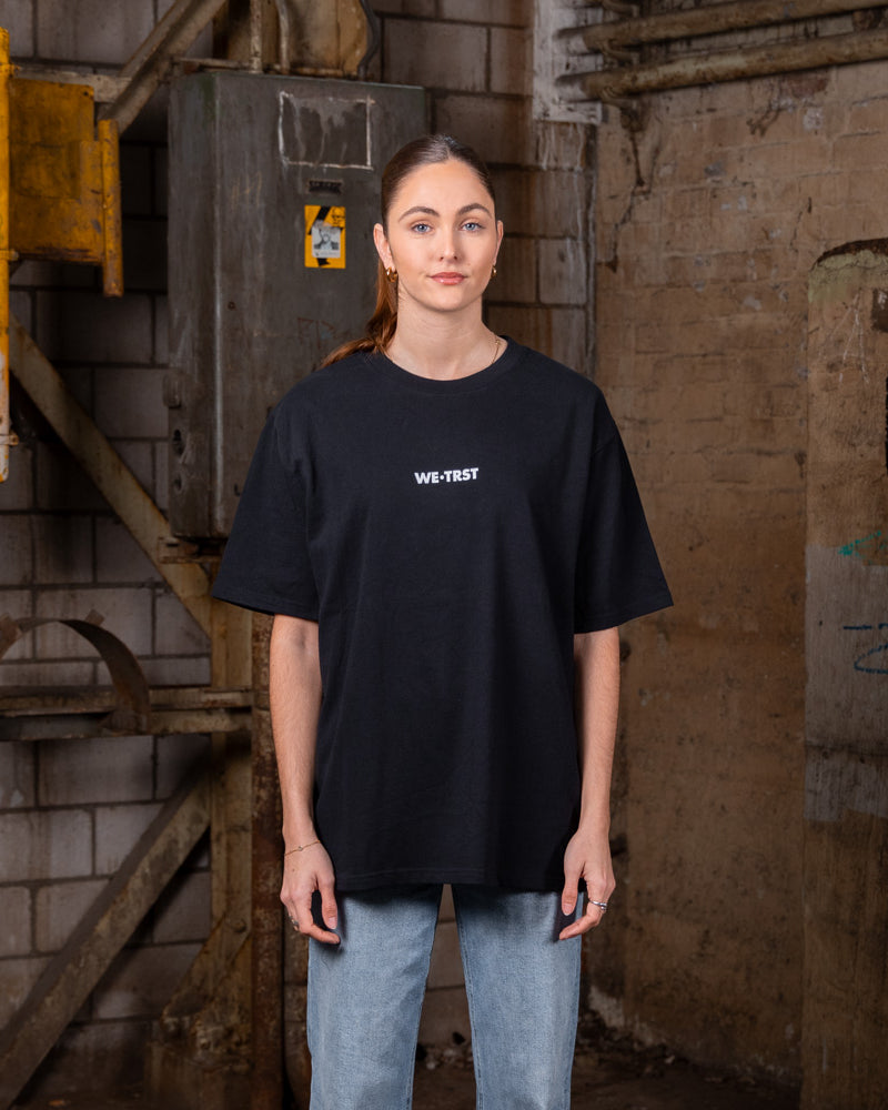 Weibliches Modell, das ein schwarzes WE•TRST-T-Shirt in Übergröße mit minimalistischem Logo trägt, posiert in einer städtischen Industrieumgebung mit freiliegenden Ziegelwänden und Metallstrukturen.