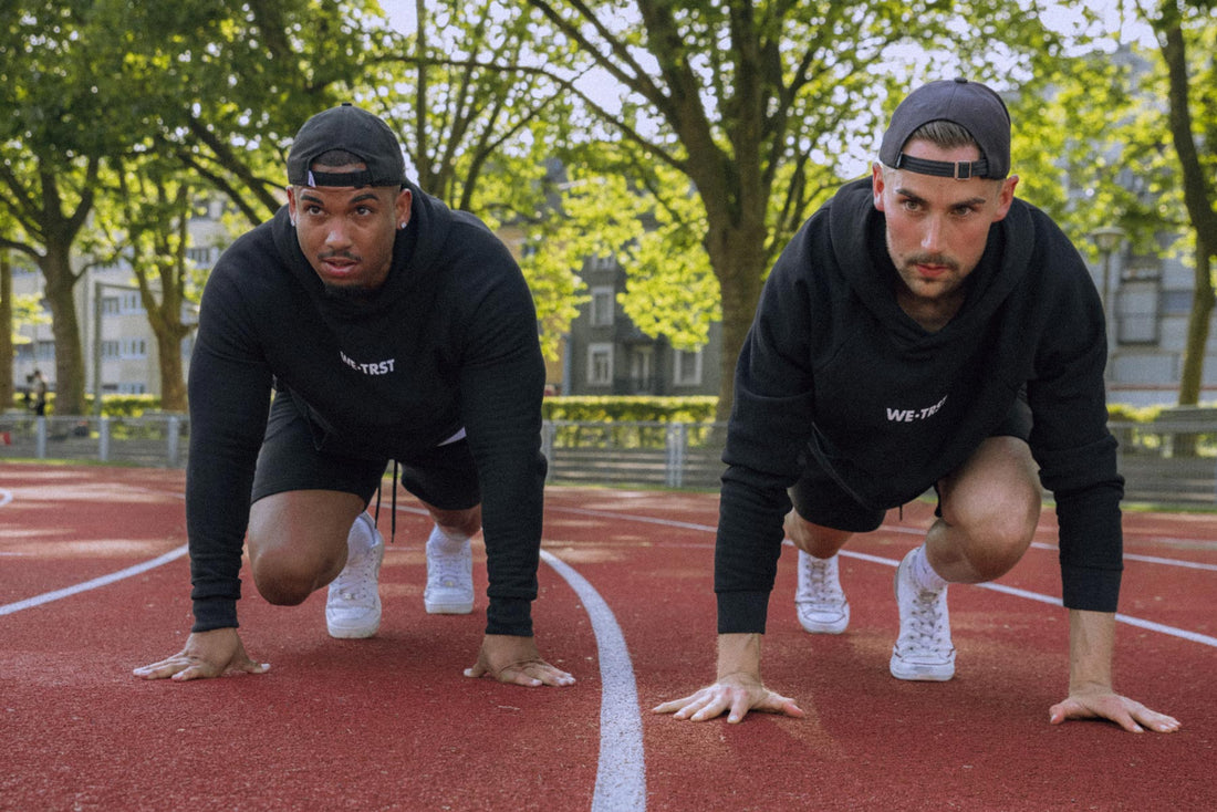 Das Bild zeigt zwei Sportler in Sprinter-Pose auf dem Boden, die beide schwarze Hoodies von WE•TRST tragen. Sie haben ihre Hände auf dem Boden platziert, während ihre Füsse in Startposition sind.