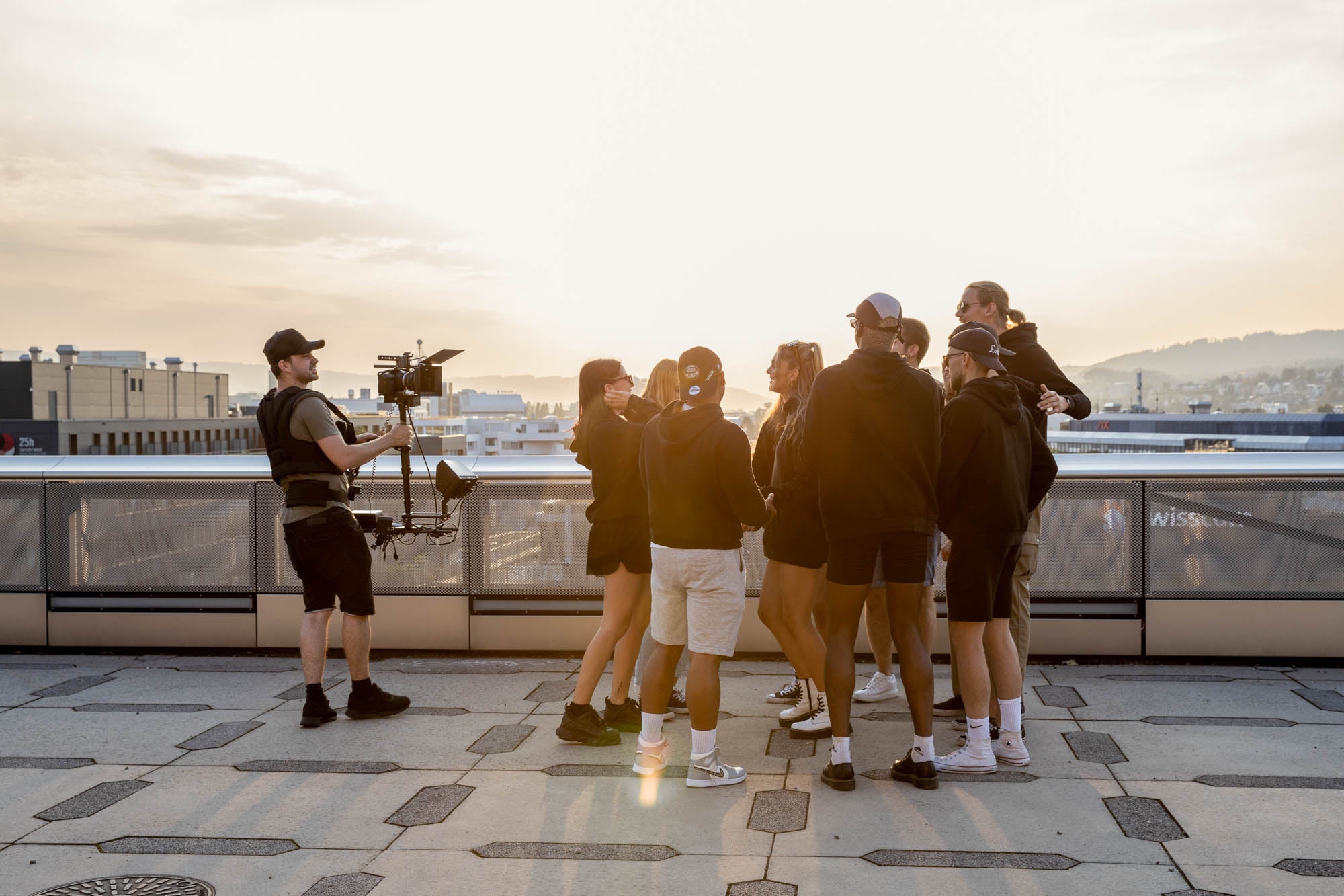 Eine Gruppe von Menschen hat auf einer Terrasse zusammen eine gute Zeit. Alle tragen schwarze Hoodies von WE•TRST. Sie lachen und unterhalten sich. Im Hintergrund ist die Sonne untergegangen und der Himmel ist in warmen Tönen von Orange und Rosa getaucht.