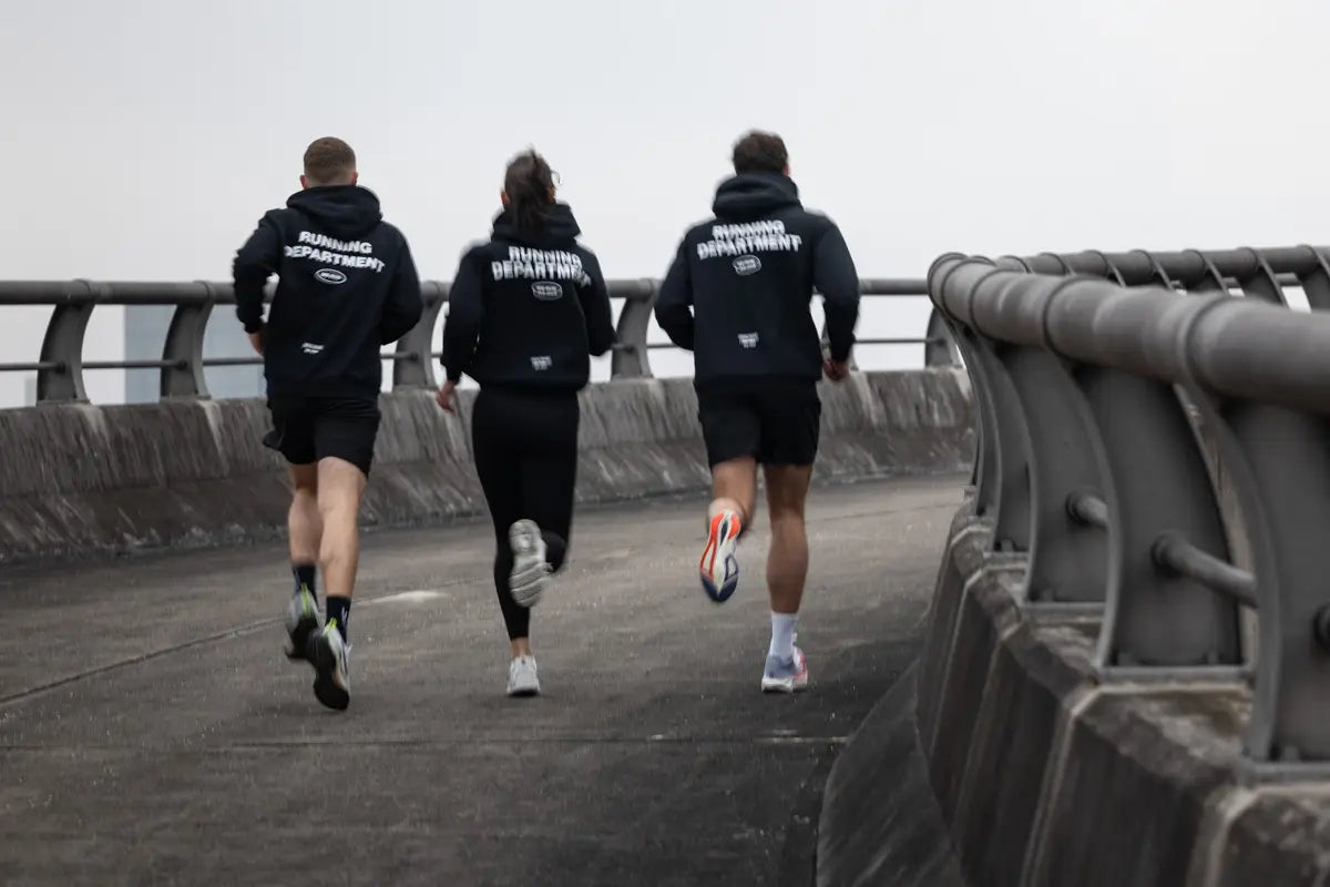 Drei Läufer:innen von WE•TRST laufen gemeinsam über eine Brücke, tragen schwarze Hoodies mit «Running Department» Backprint – Community Run, Urban Running, Laufcommunity Schweiz.