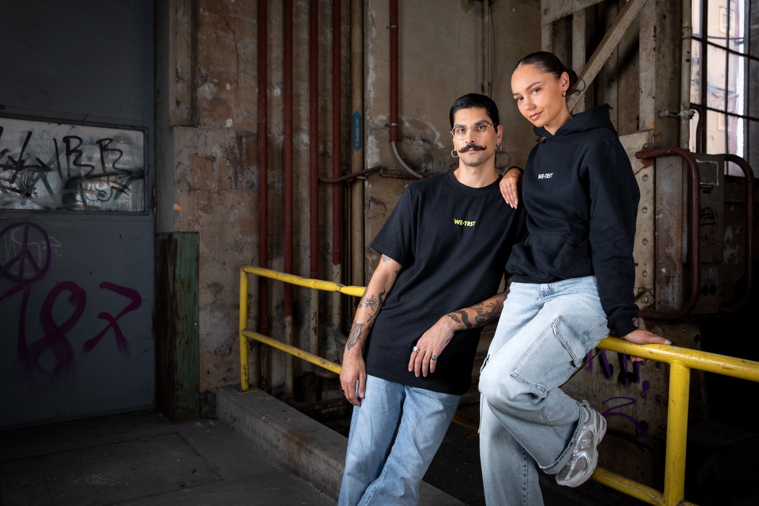 Zwei Models tragen die beiden Produkte BLACK OUT (Hoodie) und MID NIGHT (T-Shirt). Sie stehen in einer alten Fabrikhalle.