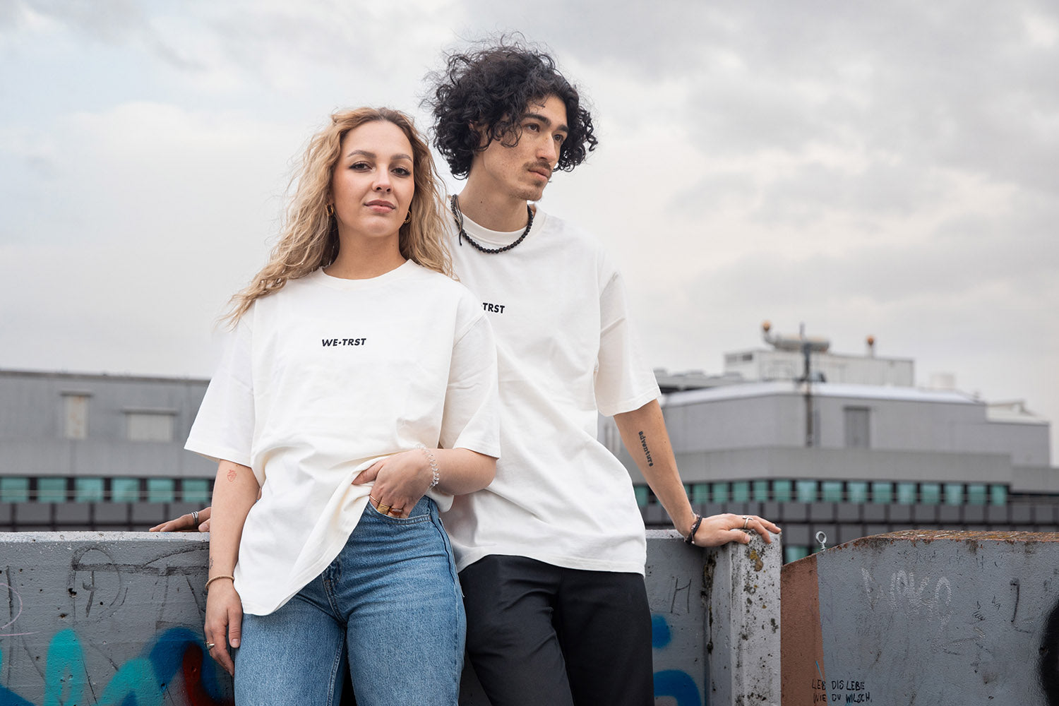 Zwei Models von WE•TRST stehen vor einem Urbanen Hintergrund mit dem neuen «Cloud Walker» T-Shirt modell. 
