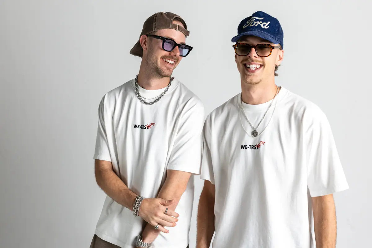 Die Dangel Twins tragen das weisse WE•TRST Dangel Twins-Shirt und lächeln in die Kamera – modernes Streetwear-Duo mit Cap, Ketten und Sonnenbrille im Studio-Setting.