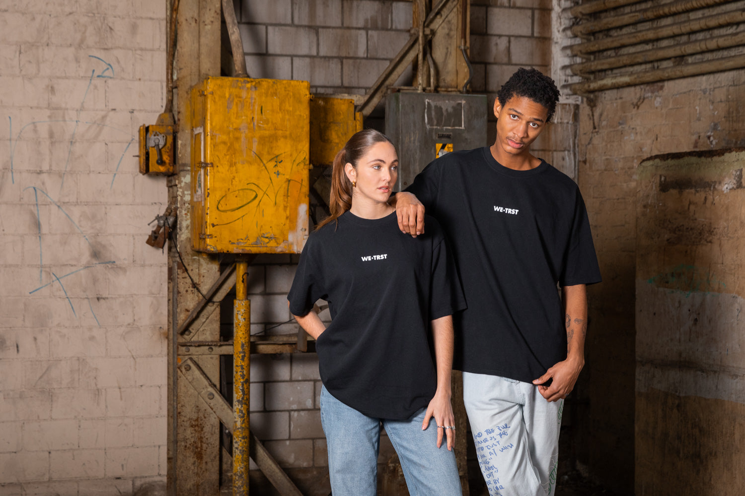 Zwei Models in lässiger Pose tragen das schwarze WE•TRST T-Shirt im Oversize-Look. Ihr entspannter Stil betont die jugendliche und lockere Ausstrahlung der Marke.