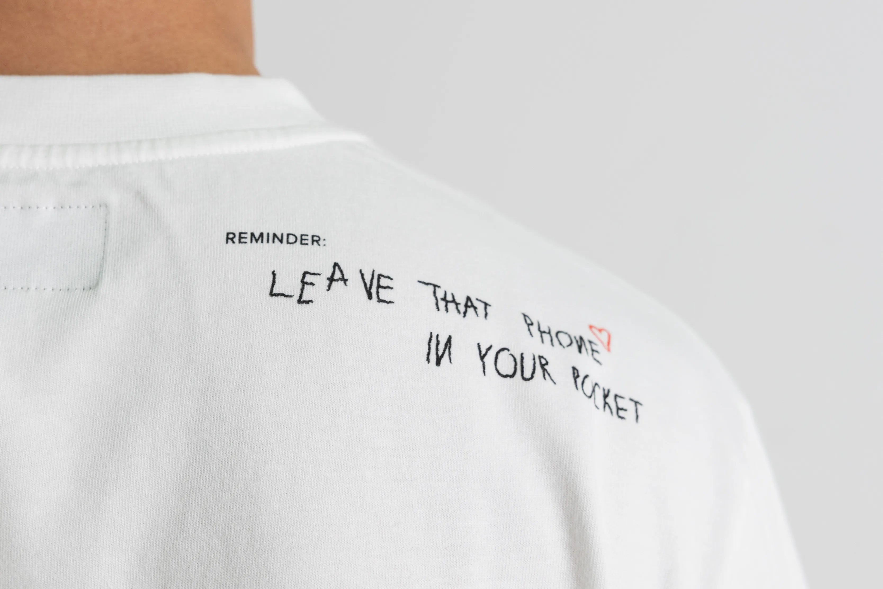 Detailaufnahme des Sticktexts «Reminder: Leave that phone in your pocket» auf dem Schulterbereich eines weissen WE•TRST x Dangel Twins T-Shirts.