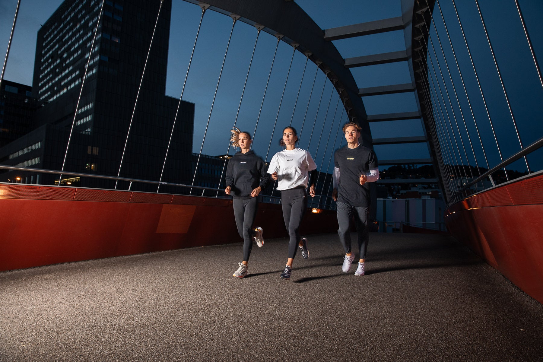 Three athletes running across a modern red bridge at night, wearing stylish activewear from WE•TRST Run Club, with illuminated city buildings in the background.