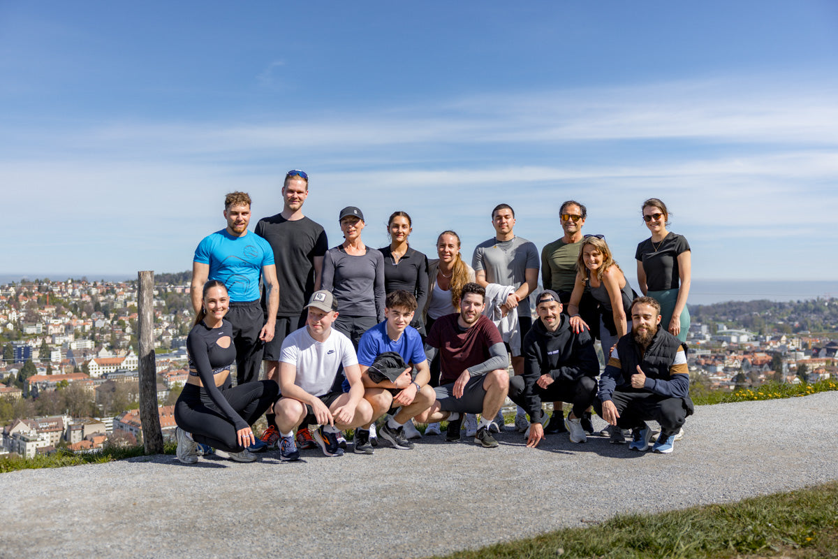 Gruppenfoto der WE•TRST Run Club-Teilnehmer, die auf einem Hügel mit Panoramablick auf eine Schweizer Stadtlandschaft unter strahlend blauem Himmel posieren und eine lebendige Gemeinschaft von Fitnessbegeisterten zeigen.