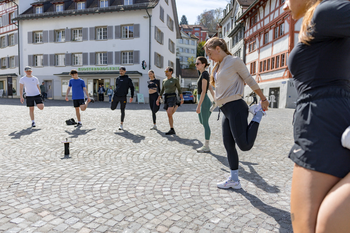 Eine Gruppe von WE•TRST Run Club-Teilnehmern wärmt sich mit Dehnungsübungen auf einem historischen Schweizer Stadtplatz auf, der von traditionellen Gebäuden und Kopfsteinpflasterstraßen umgeben ist und Gemeinschaft und Bewegung einfängt.