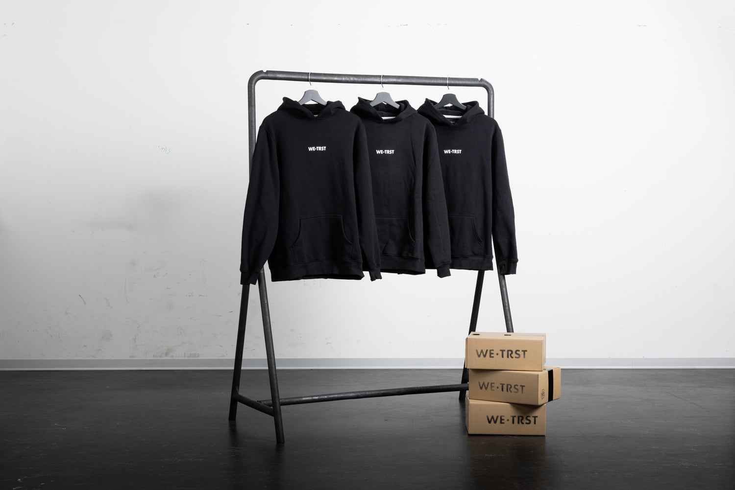 Drei schwarze Hoodies von WE•TRST an einem Kleiderständer. Darunter befinden sich drei braune Verpackungsschachteln.