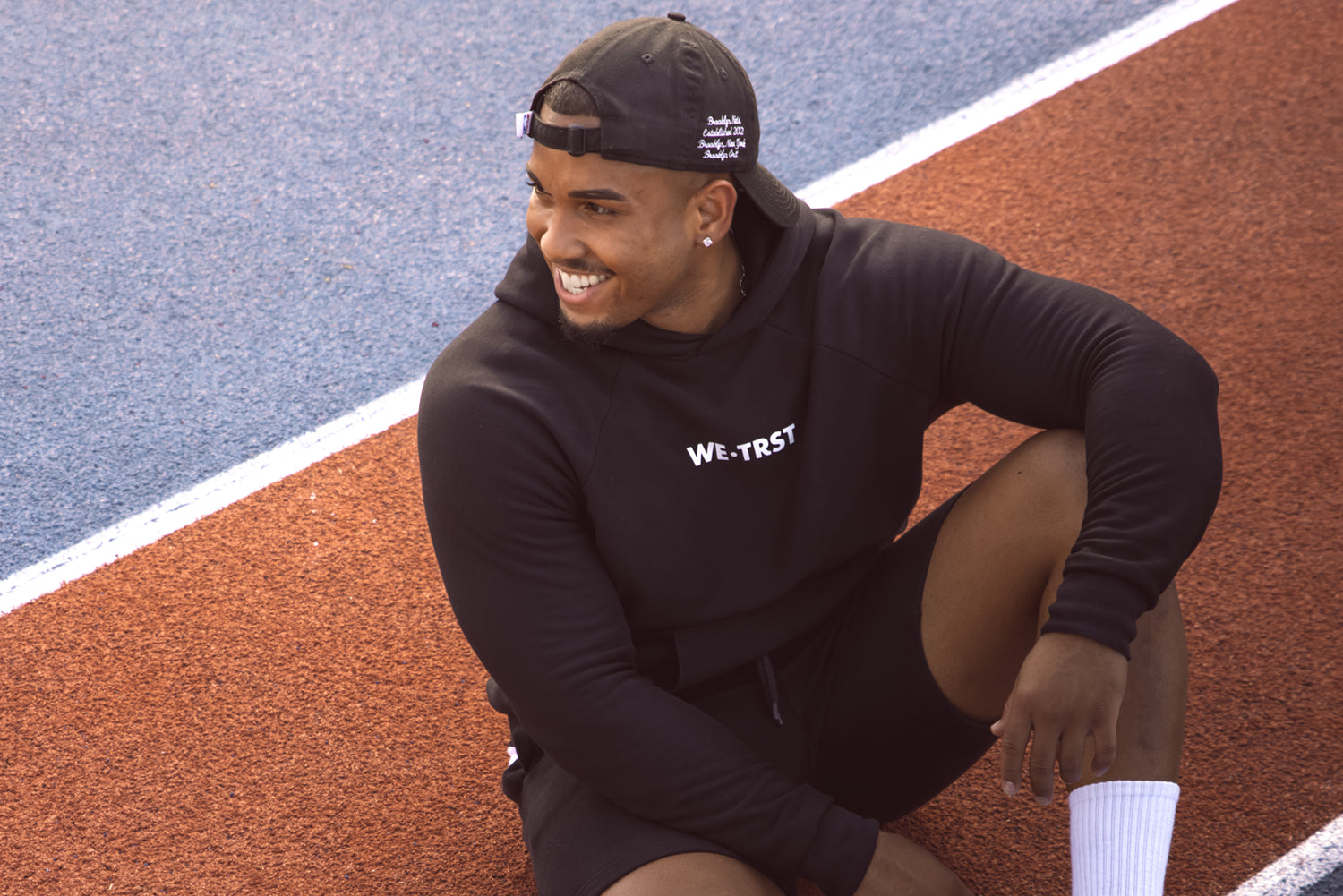 Ein junger afroamerikanischer Mann sitzt auf einem Sportplatz und trägt einen Hoodie von WE•TRST. Er strahlt ein glückliches Lächeln aus und wirkt sehr zufrieden und entspannt.