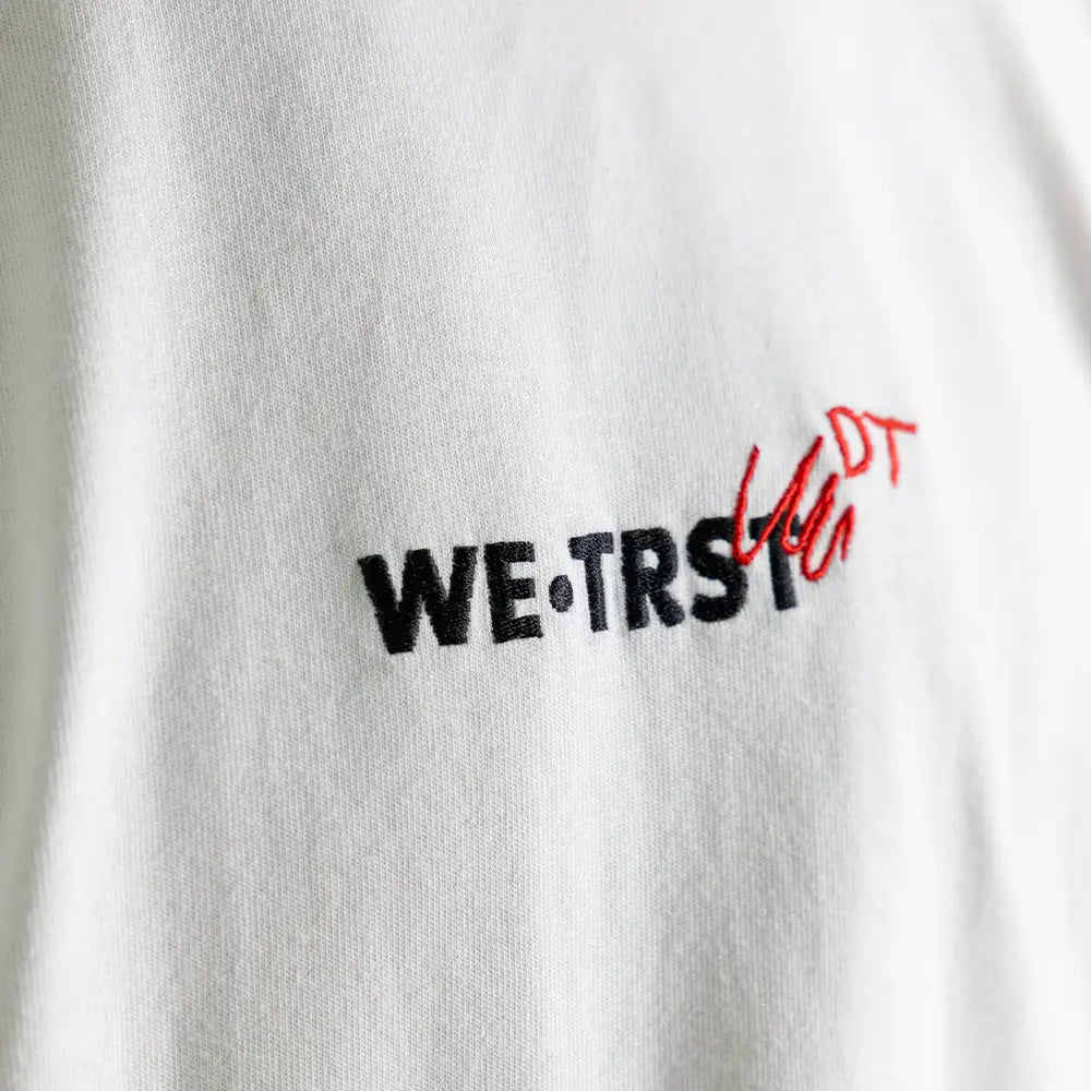 Nahaufnahme des WE•TRST Dangel Twins-Logos auf weissem Shirt – Stickerei mit rotem Doodle-Effekt.