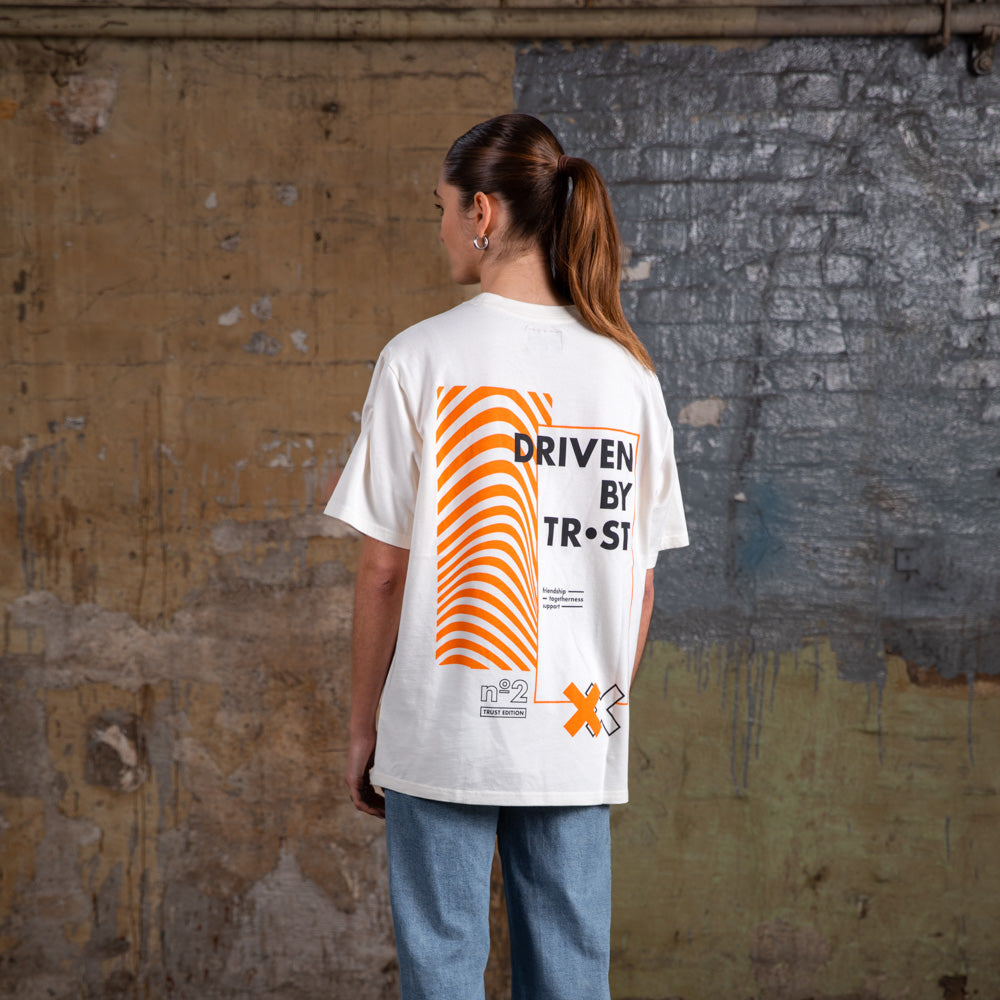 Rückenansicht eines weiblichen Models, das das WE•TRST „Driven by Trust“ T-Shirt in Übergröße trägt, wobei die auffällige orangefarbene Grafik und das typografische Design hervorgehoben werden.