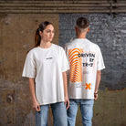 Seite an Seite zeigen männliche und weibliche Models das Vorder- und Rückseitendesign des WE•TRST „Driven by Trust“ T-Shirts in Off-White.